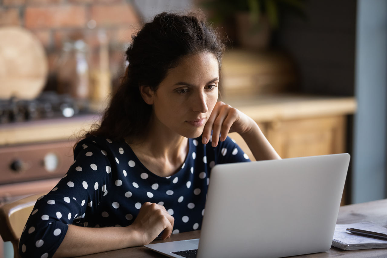 Serious young hispanic female work from home office at kitchen read e book document on laptop pc ponder on project task. Focused lady remote student look on computer screen watch web tutorial online