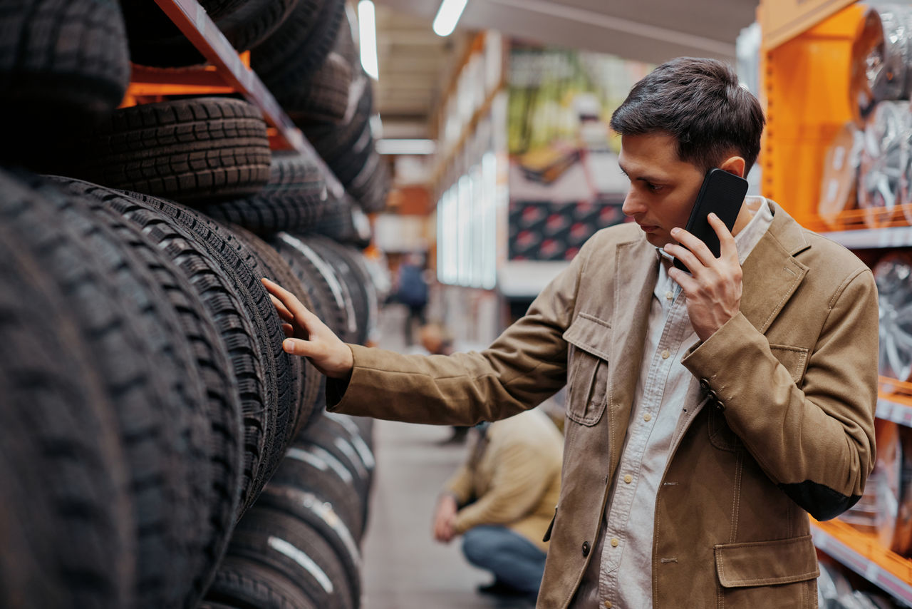 man customer choosing new tires and consulting with somebody by phone in the auto shop with car goods.