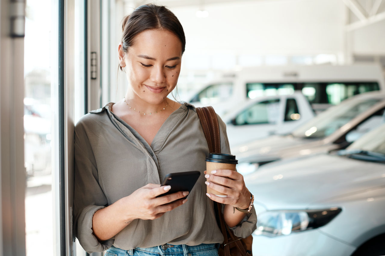 woman looking at phone at car dealership while drinking coffee