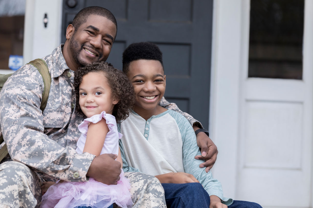 Happy military soldier is excited and relieved to be reunited with his preschool age daughter and preteen son. They are sitting on the front porch of their home.