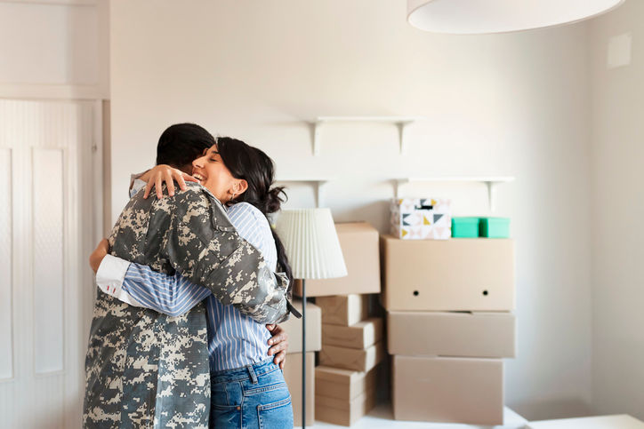 Wife embraces her army veteran husband in camouflage clothing while they moving into the new house. First time home buyers celebrate and hugging new house purchase on moving day, laughing standing among boxes in own flat house
