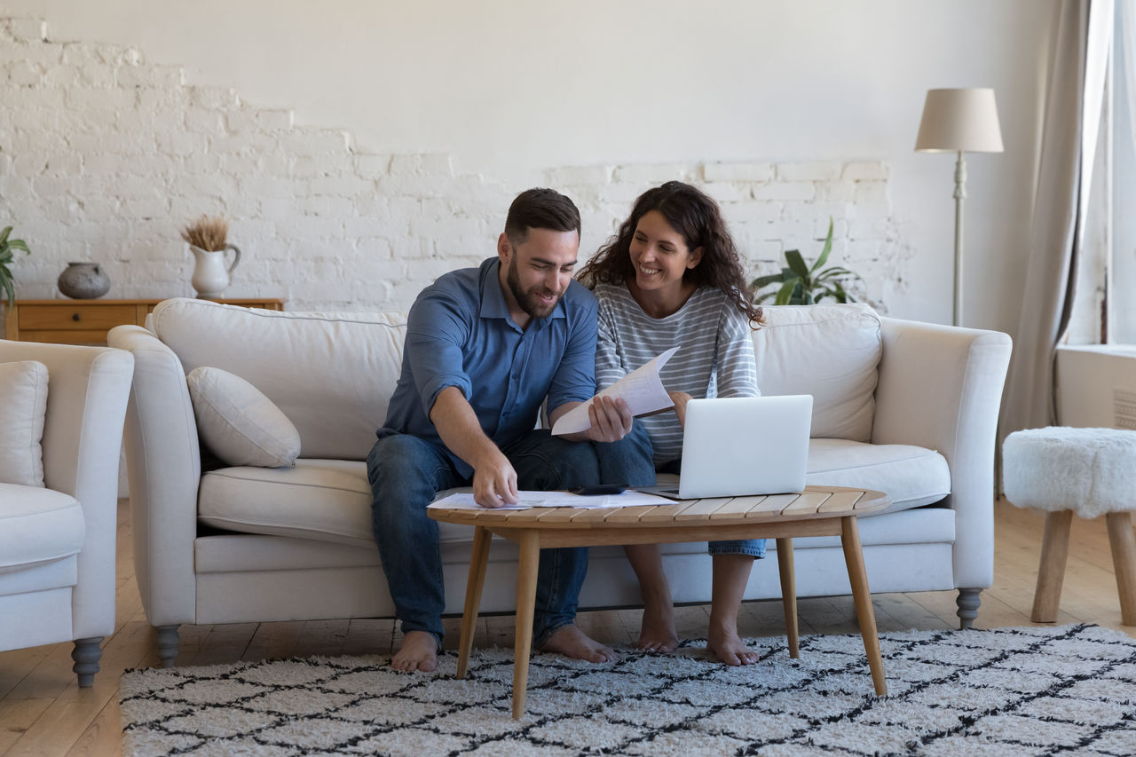Millennial wife and husband sorting out bank papers, check invoices, paying bills via e-bank on laptop sit on sofa smile feel satisfied. Enough money, payments, profit, financial management concept