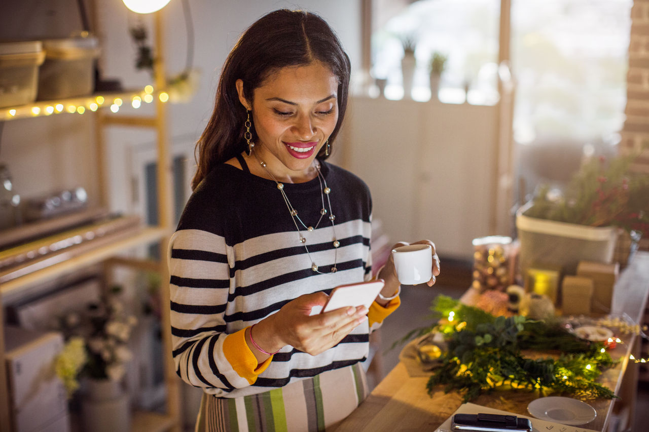 Working woman at gift shop. She wearing casual clothing, checking online shop with phone to receive new orders from customer