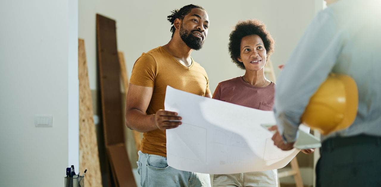 Happy African American couple analyzing blueprints while communicating with a building contractor in the apartment.