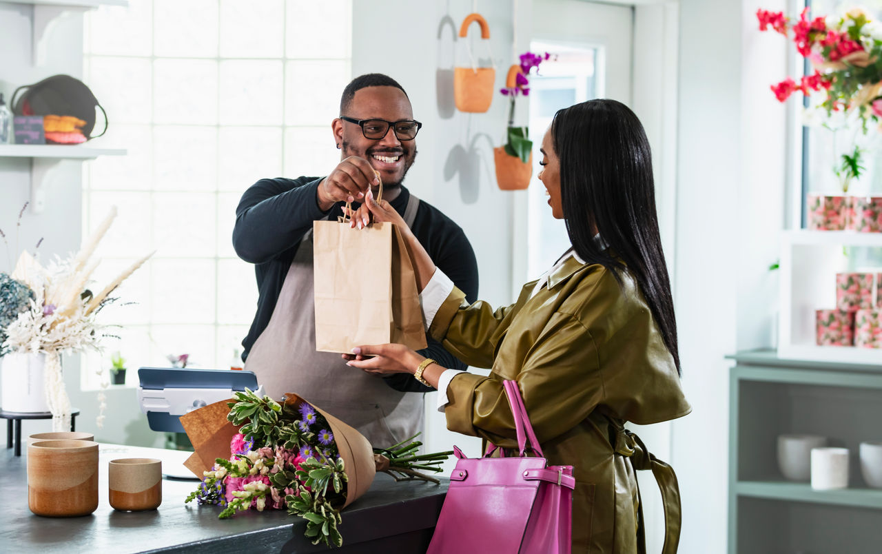 A mid adult African-American man, in his 30s, working at the checkout counter of a gift and flower shop, helping a customer make a purchase. The customer is a young African-American woman in her 20s.