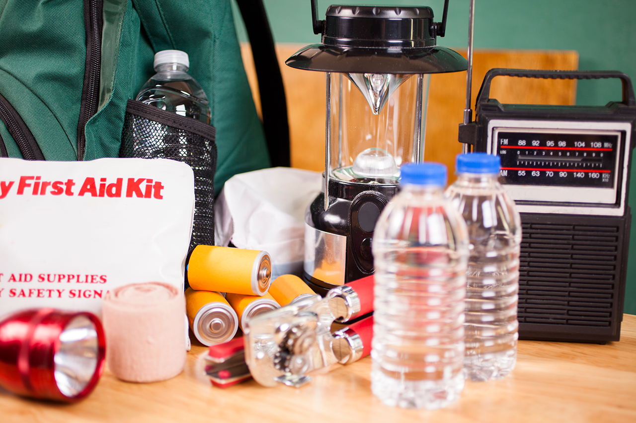 Emergency preparedness supplies. A large pile of supplies to be used in case of a natural disaster (hurricane, flood, earthquake, etc.) including: flashlight, backpack, batteries, water bottles, first aid kit, lantern, radio, can opener, mask.  No people.