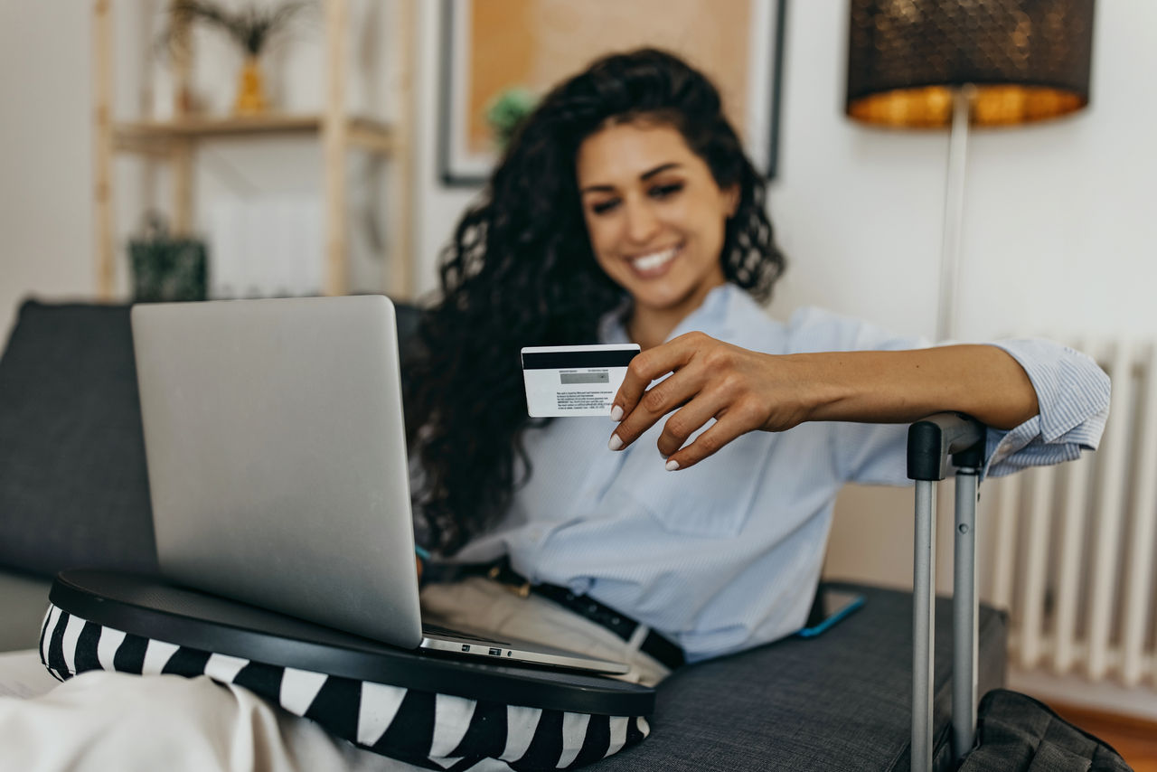 Young woman sitting at home with her bags packed, using laptop and holding credit card to pay for holiday reservation online