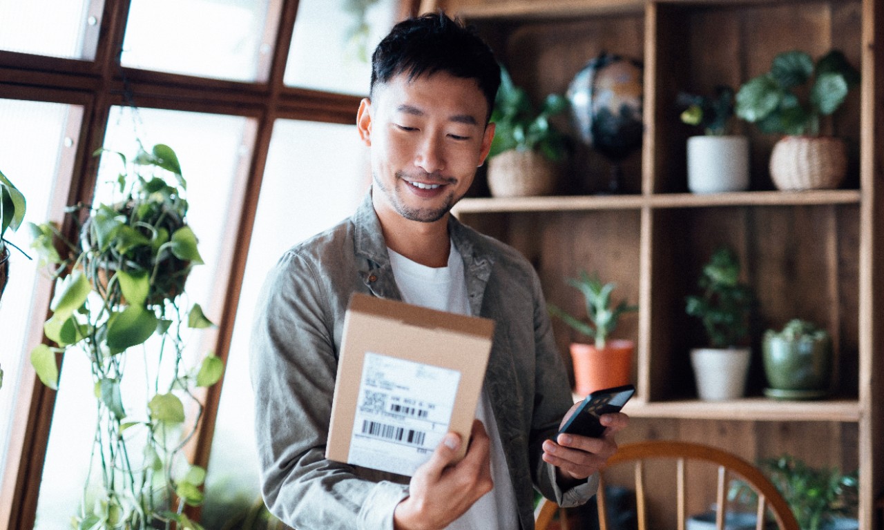 Smiling young Asian man with smartphone, receiving delivered packages from online purchases at home, can't wait to unbox the purchases. Online shopping, enjoyable customer shopping experience