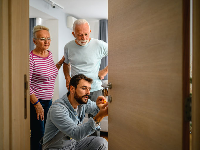 Man fixing a lock on the door while a senio coule is standing next to him.
