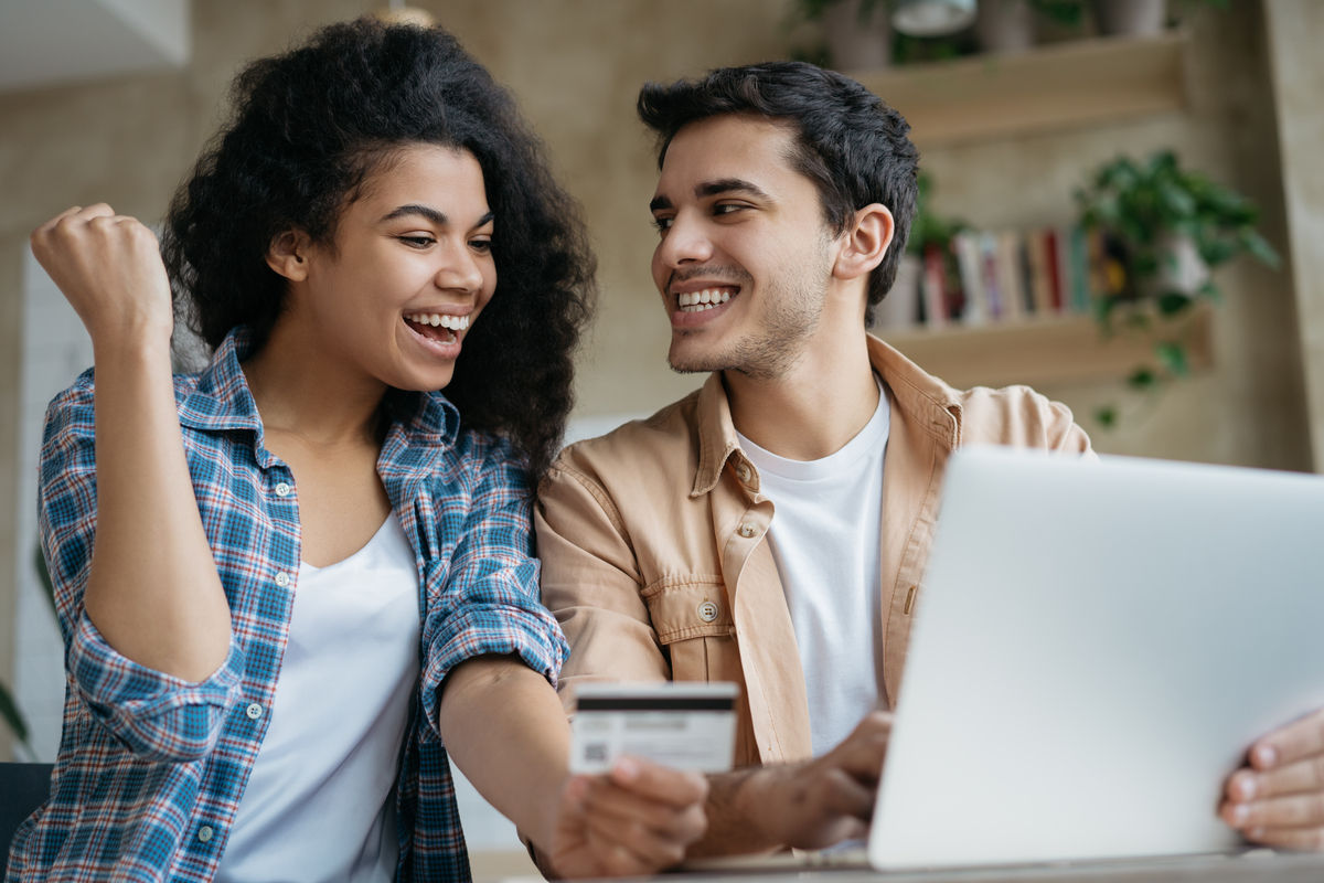 Young happy couple shopping online, sitting at home. Beautiful African American woman holding credit card, attractive man booking tickets, ordering food on website