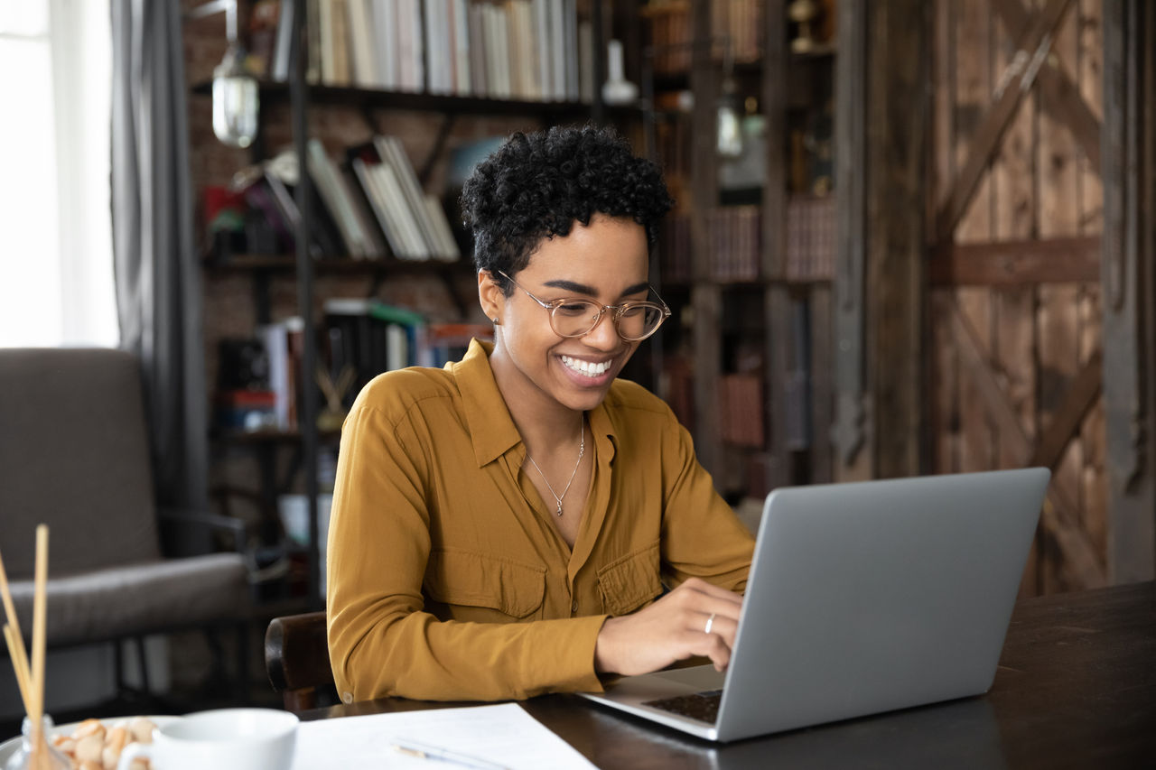 African woman sit at desk working on laptop, student studying makes assignment looks happy, feels satisfied. Workflow use modern tech, chat on-line, share messages with friends in social media concept