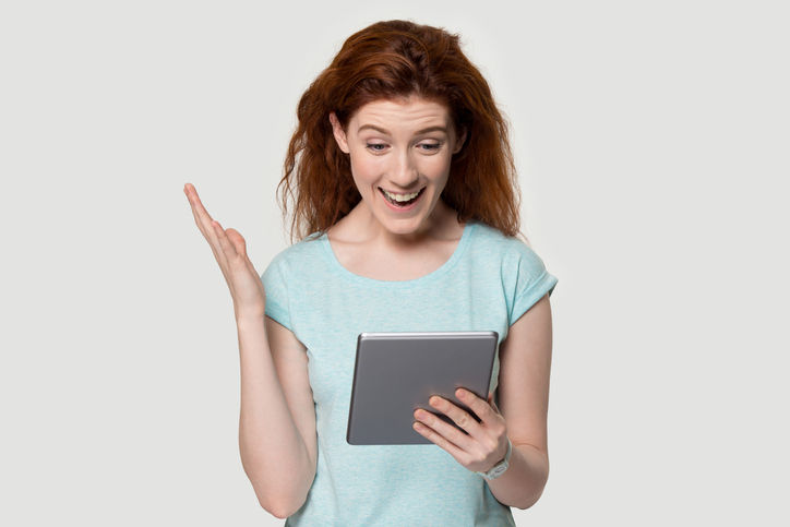 Excited young red-haired woman looking at tablet screen, feeling amazed, surprised by cool software update, limited product available, winner emotions, prize receiving, isolated on studio background.