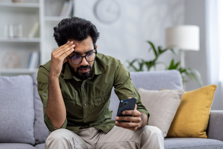 Upset and sad man sitting on sofa at home in living room, hindu man reading bad news online from phone, depressed and sad man.