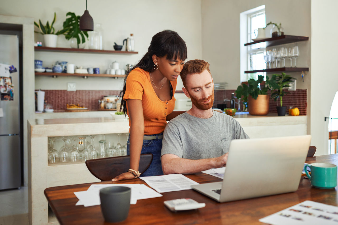 Shot of a man sitting with his laptop and paperwork while his girlfriend stands behind hi