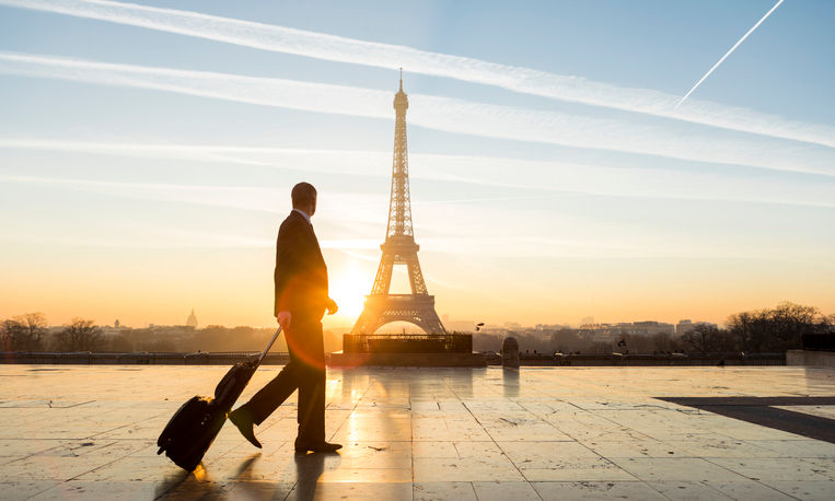 A beautiful sunrise takes place in Paris behind the iconic Eiffel Tower and a young businessman walks with his business luggage and contemplates the iconic monument. Concept of worldwide business travel, mobility and business confidence.