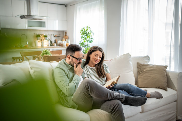 Happy Caucasian couple relaxing at home, sitting on the sofa in the living room and enjoying reading a book together