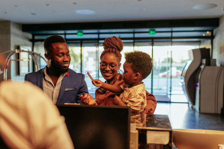 A happy African American family is checking into the hotel. The child is handing it's father the room key it just got from the receptionist.