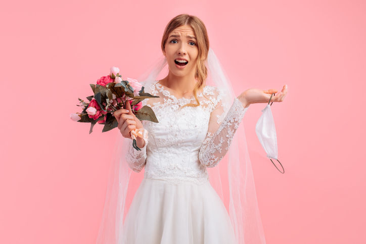 Beautiful bride in a wedding dress with a medical protective mask in her hands, on a pink background. Quarantine, wedding, coronavirus