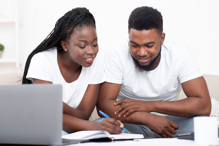 Young african american couple planning budget together at home, writing notes to notebook, creating checklist for shopping or guest list for wedding