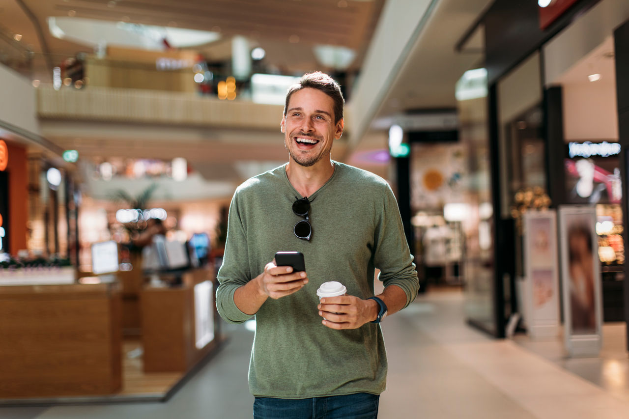 Happy man texting and drinking coffee in shopping mall