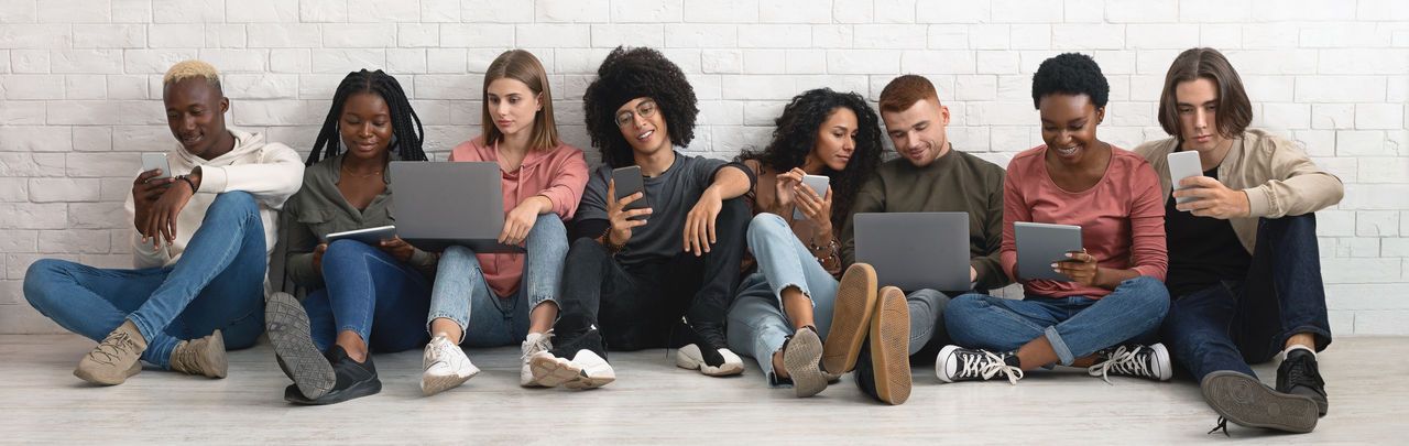Happy multiethnic millennials in casual sitting on floor by white brick wall and using various gadgets laptops, digital tablets, cellphones while spending time together, chatting, panorama