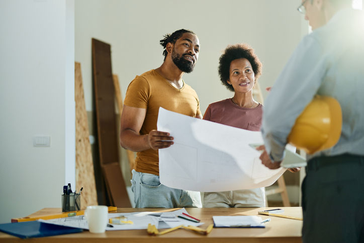 Happy African American couple analyzing blueprints while communicating with a building contractor in the apartment.