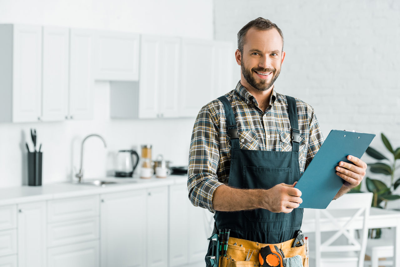 smiling handsome plumber holding clipboard and looking at camera in kitchen