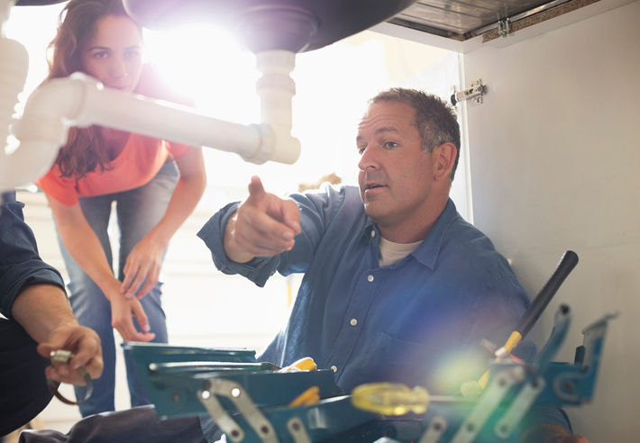 white male plumber sitting under sink inspecting pipe in white couple's home