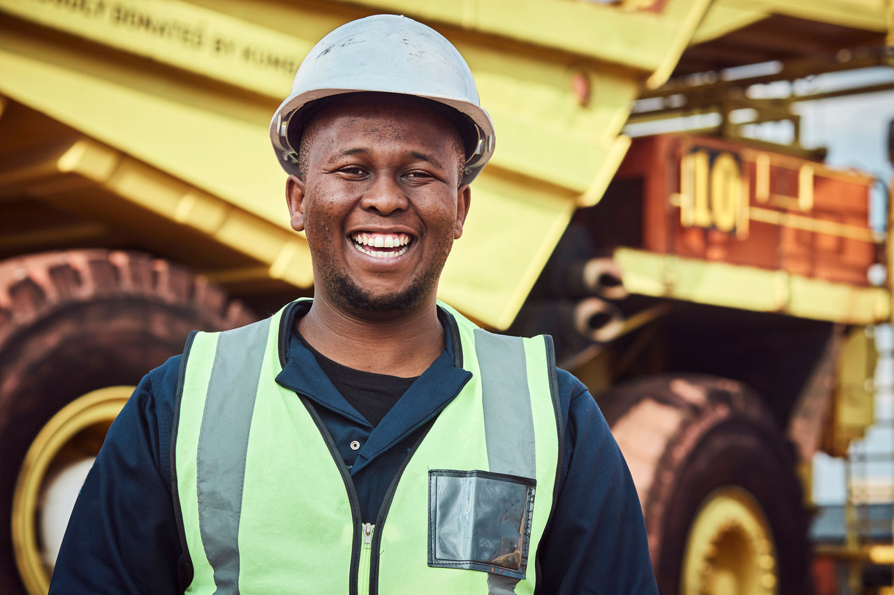A young African mine worker is standing with a smile in front of a large haul dump truck wearing his personal protective wear