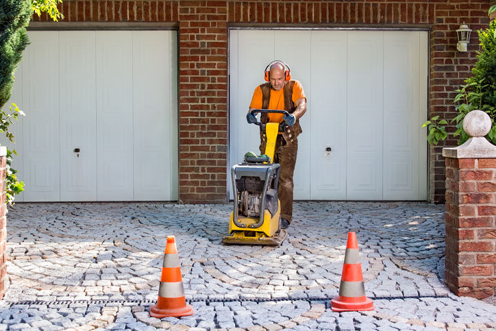 Builder or contractor laying new paving bricks in a house driveway using a mechanical compacter for compaction of the cement pavers.