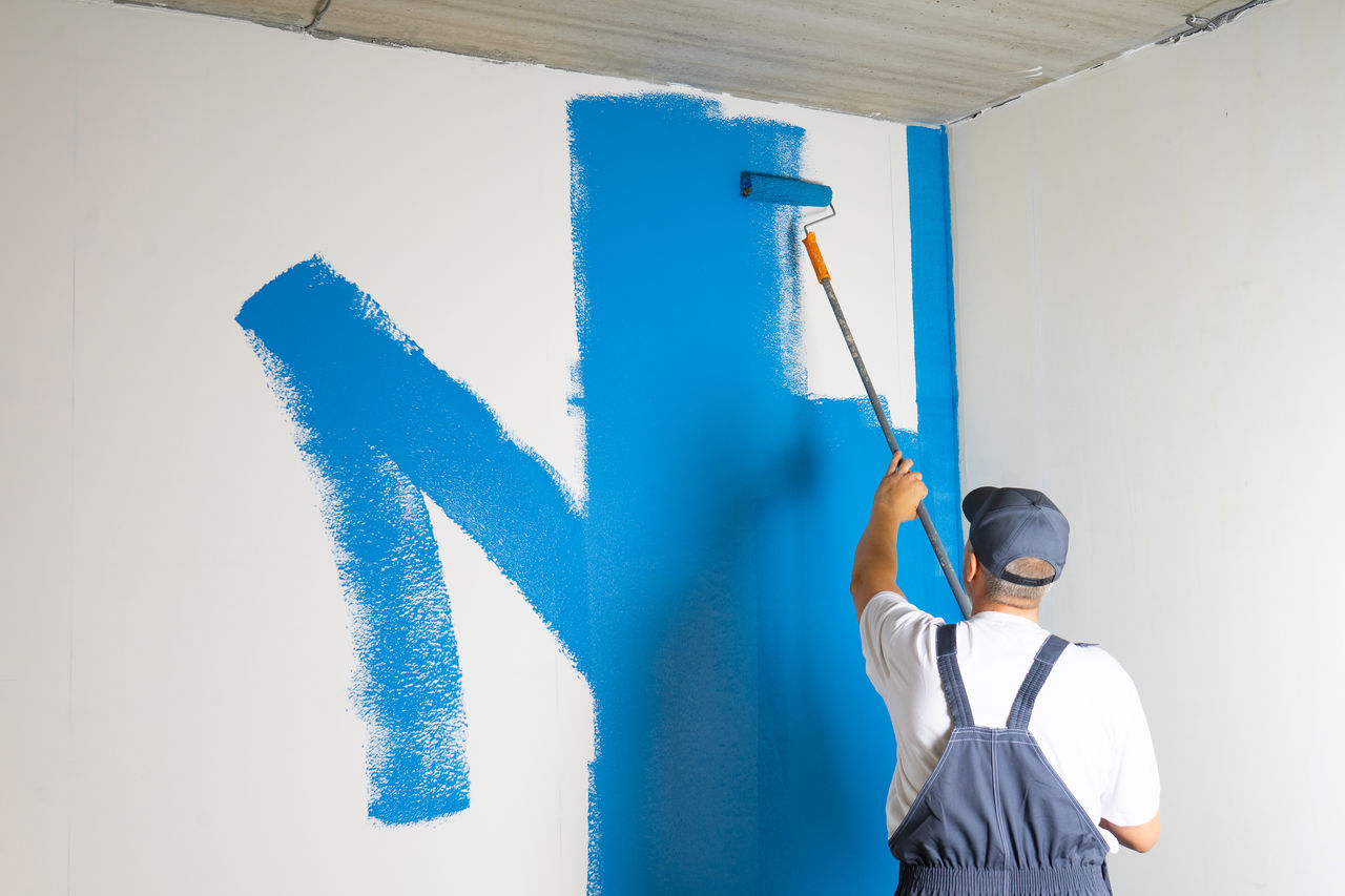 Male painter painting a white wall with a roller in blue color renovations or construction concept