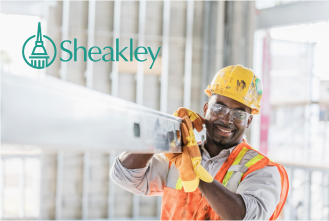 A mid adult African-American man in his 30s wearing a hard hat, protective goggles, a safety vest and work gloves, carrying a piece of construction material into the structure being built. The construction worker is smiling, looking at the camera.
