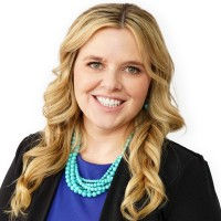 Carly Buchanan's head shot smiling woman with long blond hair in a blue shirt and black business jacket and large teal beaded necklace