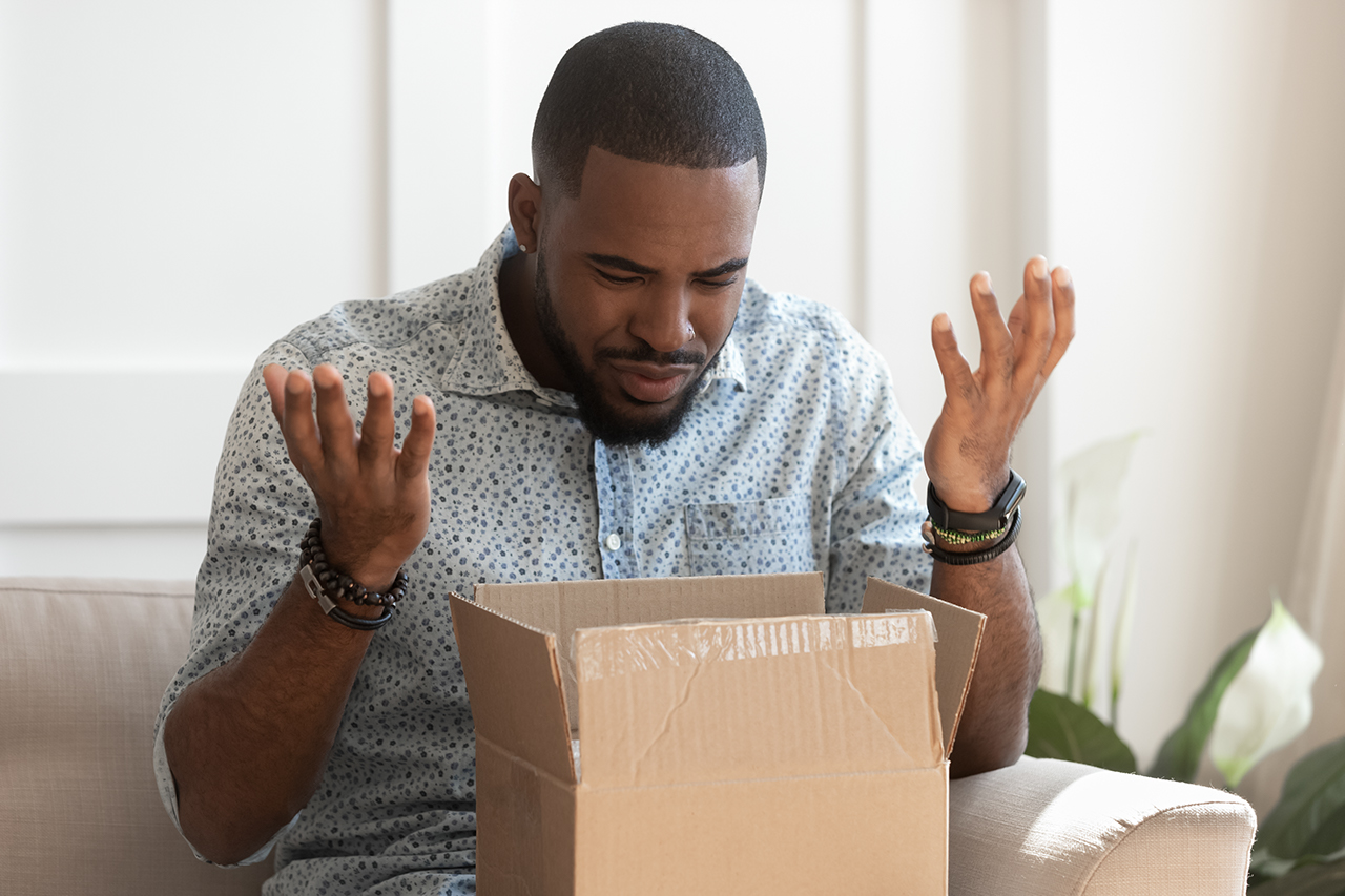 Frustrated african American millennial male buyer unpack unbox cardboard box delivery package disappointed with product quality, confused biracial man shop online open box get wrong order