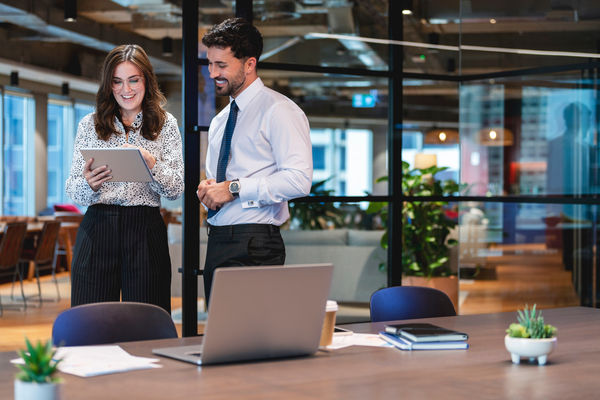 Business Man and woman standing working together on a digital tablet. They are both well dressed standing in a modern office with a laptop and paperwork on the desk. They are happy and smiling