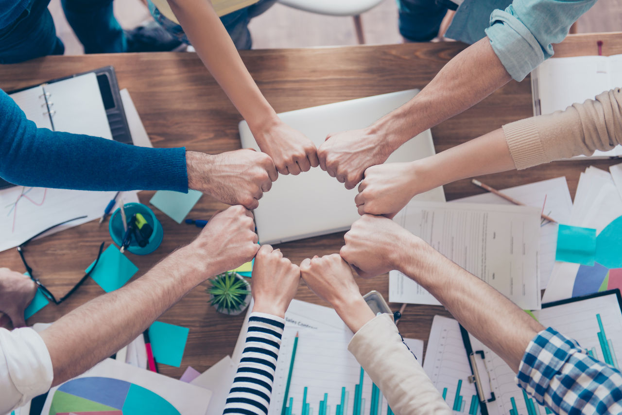 Close up cropped photo of partners putting their fists together in a circle on top of the table with work stuff. Trust, friendship, unity, cooperation concept