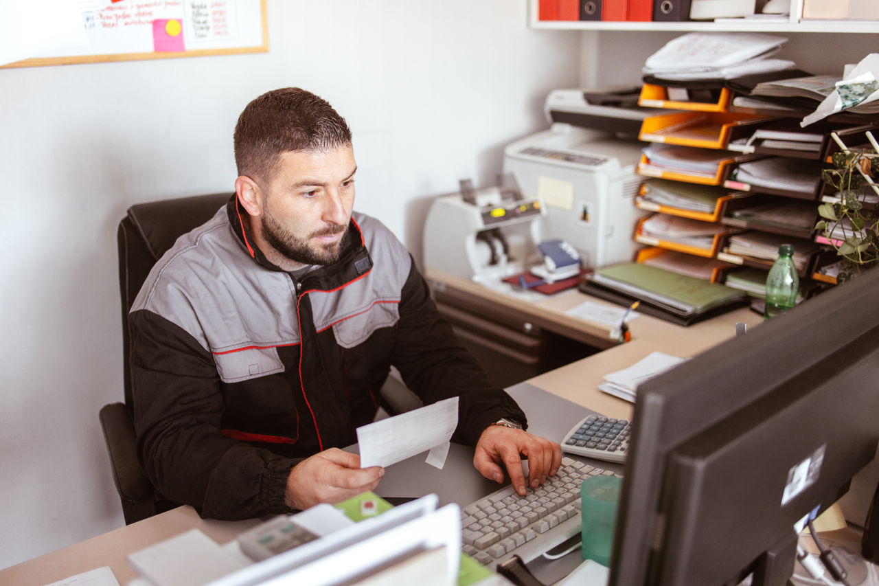 The car service technician writes notes while working on the computer in the offices and checks the invoice issued by the client.