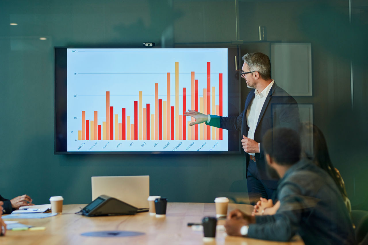 Confident mature businessman presenting financial statistics to diverse group of colleagues during meeting in modern boardroom. Professionals discussing data and graphs on large screen. Concept of corporate teamwork and leadership.