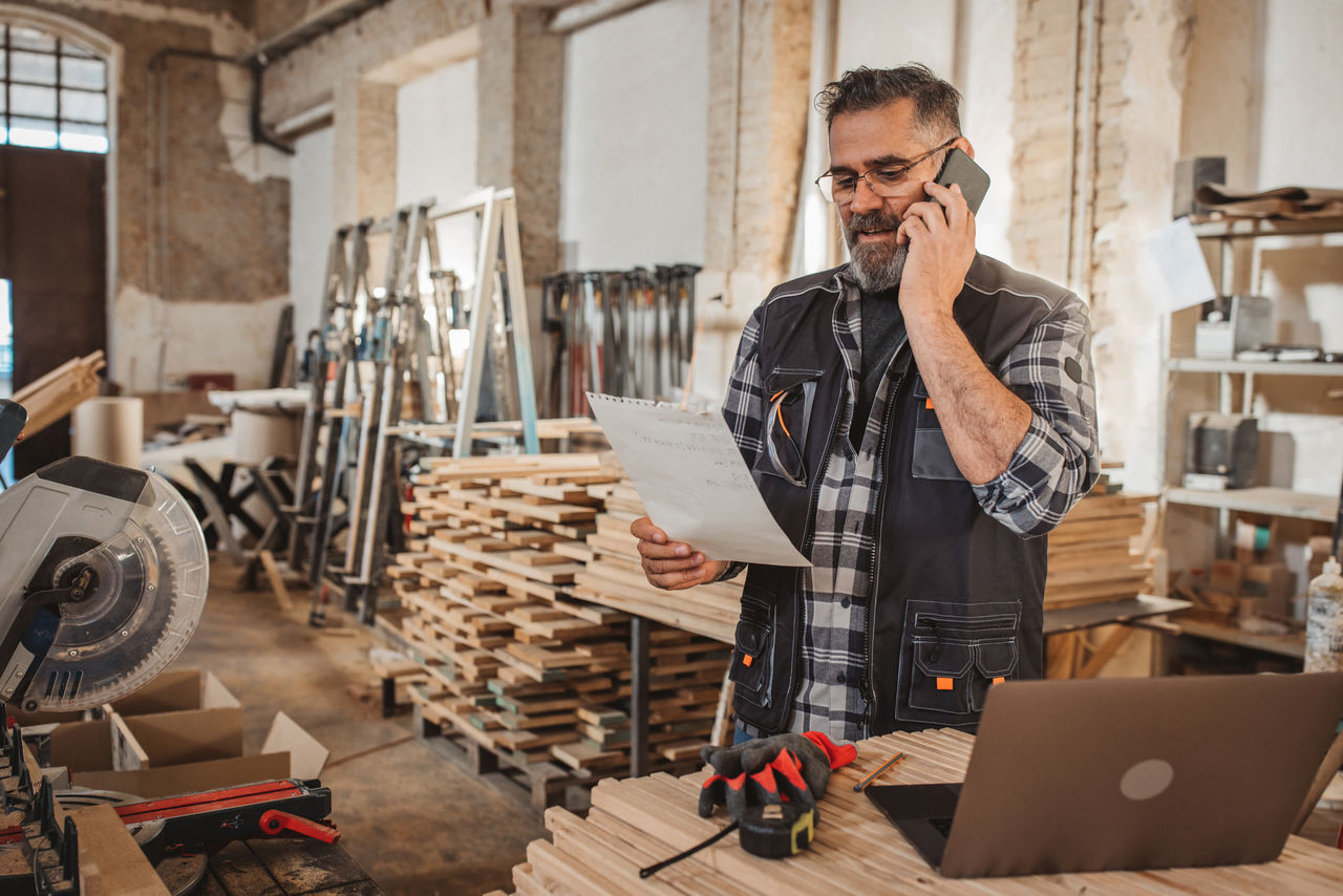 Carpenter in wood workshop using laptop and working on project, online shopping or banking.