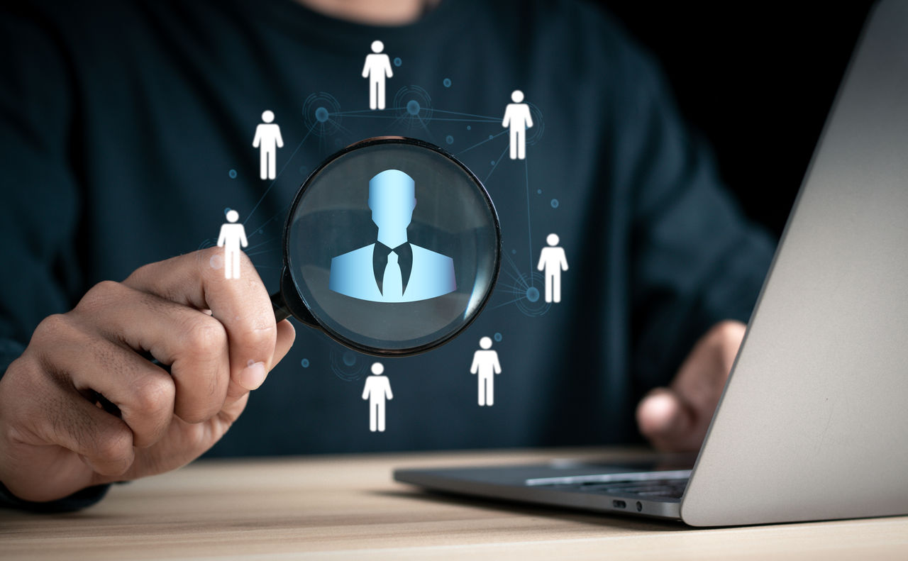 Businessman using magnifying glass to look at personnel icon on virtual social network interface. Personal network structure. Concepts of human resource management and recruitment.