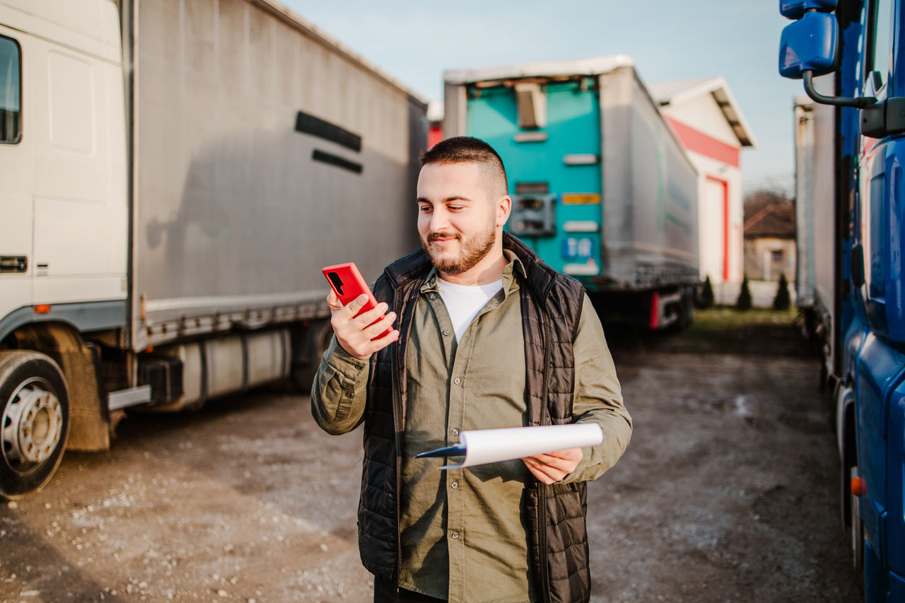 Young handsome truck driver standing in front of his truck and using smart phone for communication.