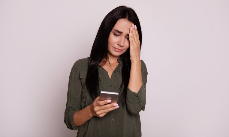Close-up photo of a gorgeous girl with long raven black hair, who is reading something on her smartphone with a sad face expression and holding her head with her fingers.