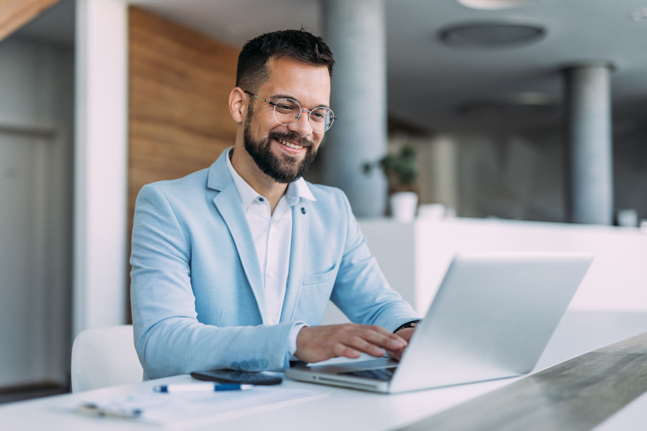 Shot of a businessman sitting on desk in modern office and working on laptop. Elegant handsome businessman sitting in his office and using laptop.