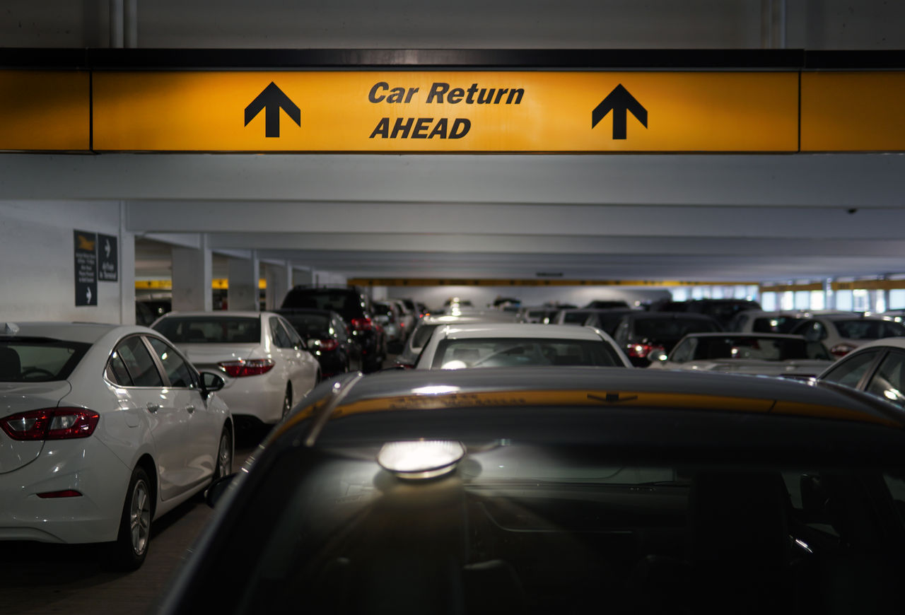 yellow and black cars in a row from backside, a yellow sign board with arrows and letters showing the way for car return ahead