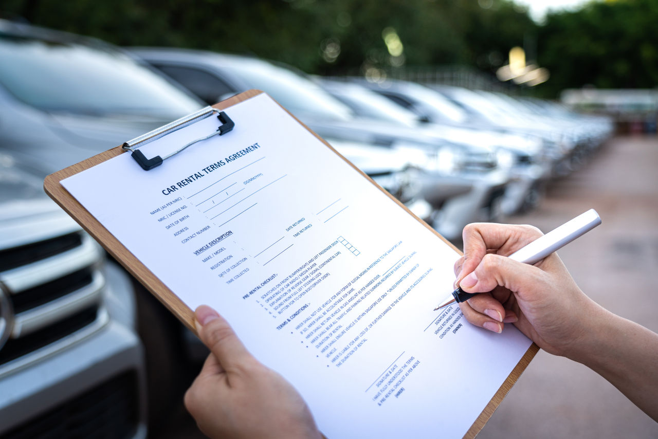 Action of a customer is signing on the agreement term of car rental service. Close-up and selective focus a human's hand with blurred background of cars in row. Business and transportation concept.