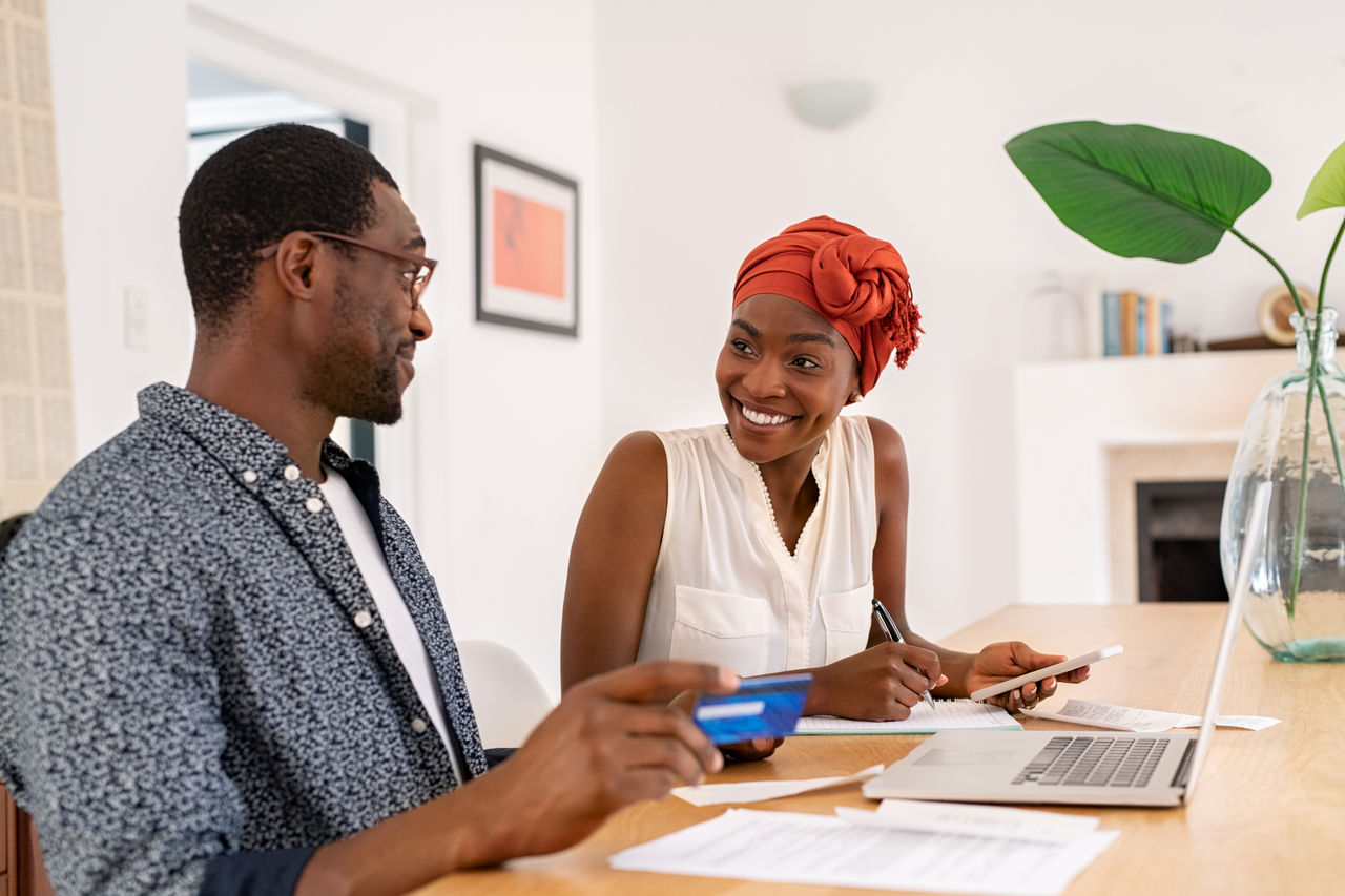 Mature smiling couple sitting and managing expenses at home. Happy mid black man and woman with traditional turban paying bills and managing budget. Middle aged african american couple checking accountancy and bills while holding credit card.