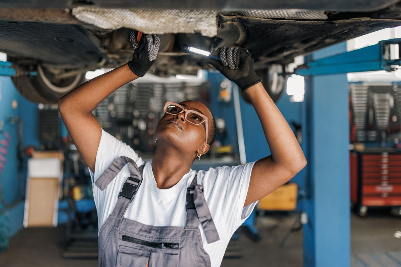 A young African American female mechanic is standing under a car and examining the damage on the broken parts with a serious look on her face.