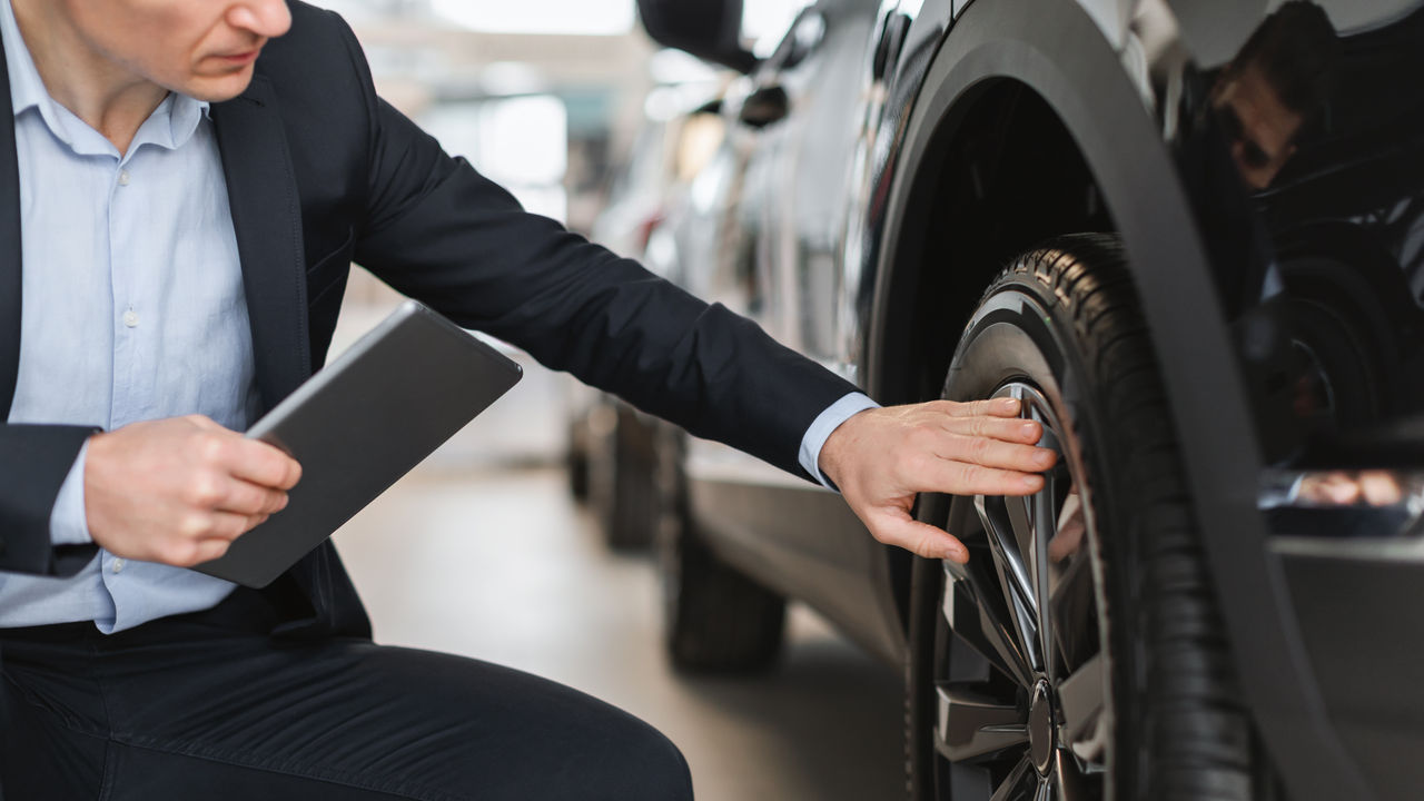 Unrecognizable auto salesman sitting near brand new car, checking wheel and tire at modern dealership, closeup. Young insurance agent examining automobile at showroom store, panorama