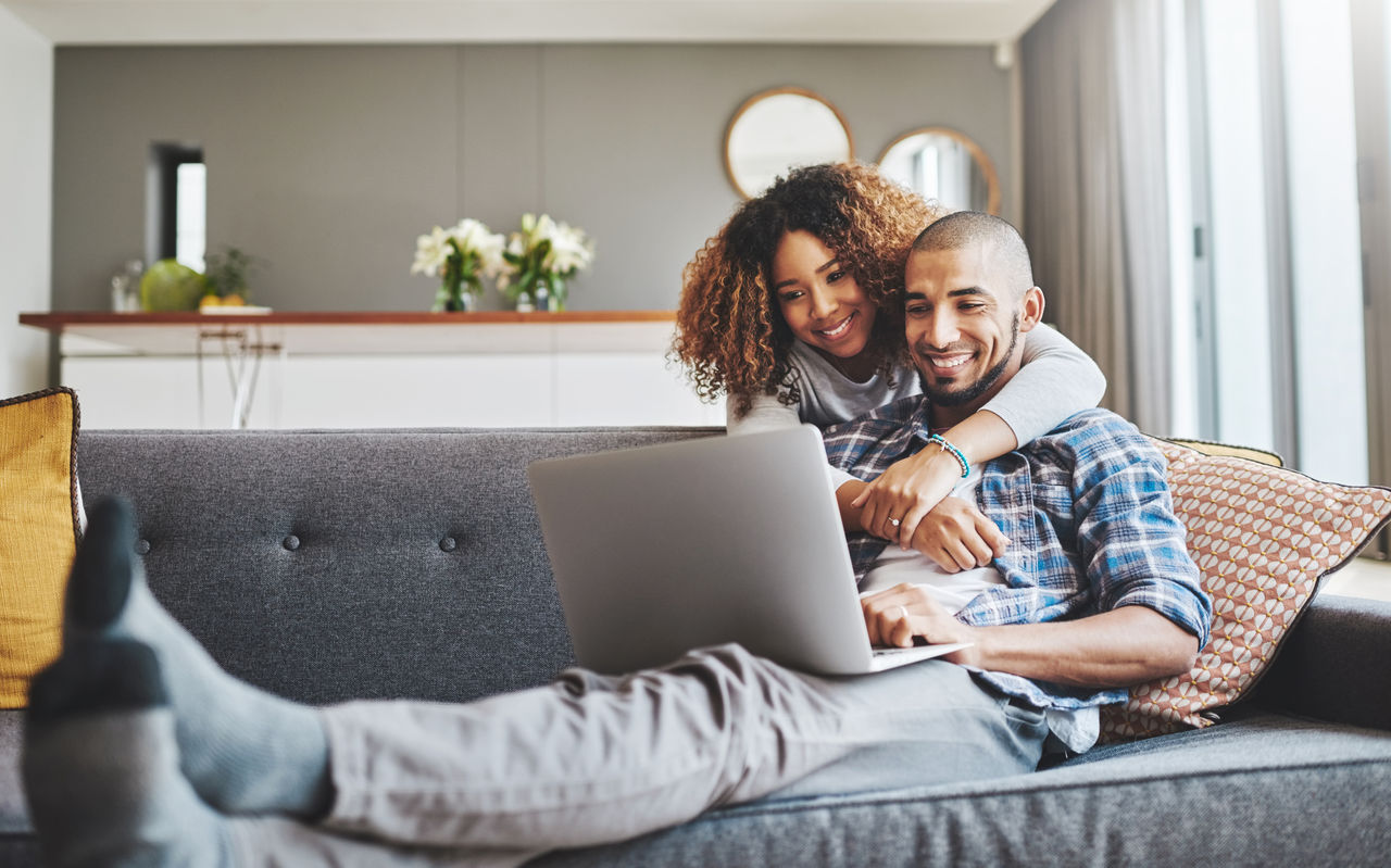 Shot of a young woman hugging her husband while he uses a laptop on the sofa at home