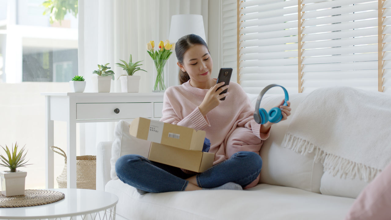 Young asia people happy teen girl smile unbox open gift new headphone buy order from online store shop take photo shoot camera show post social media app blog vlog share sit relax at home sofa couch.